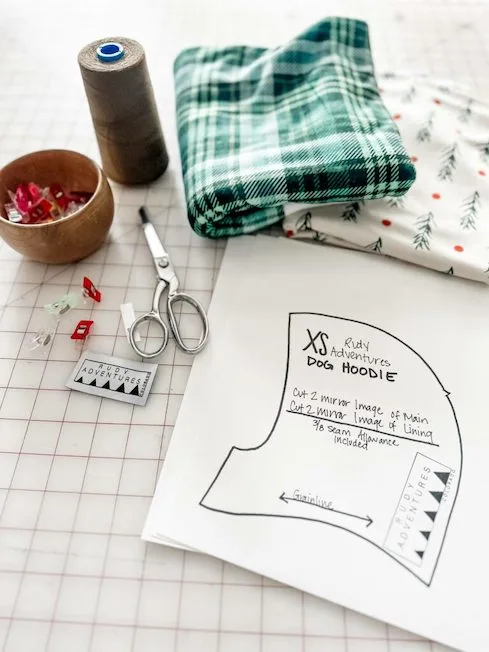 Cut-out pieces of square paper rest on a grid surface near a pair of scissors, a spool of thread and sewing pins. A folded piece of green, black and white plaid fabric lays on a white piece of fabric with red dots and evergreen tree leaves.