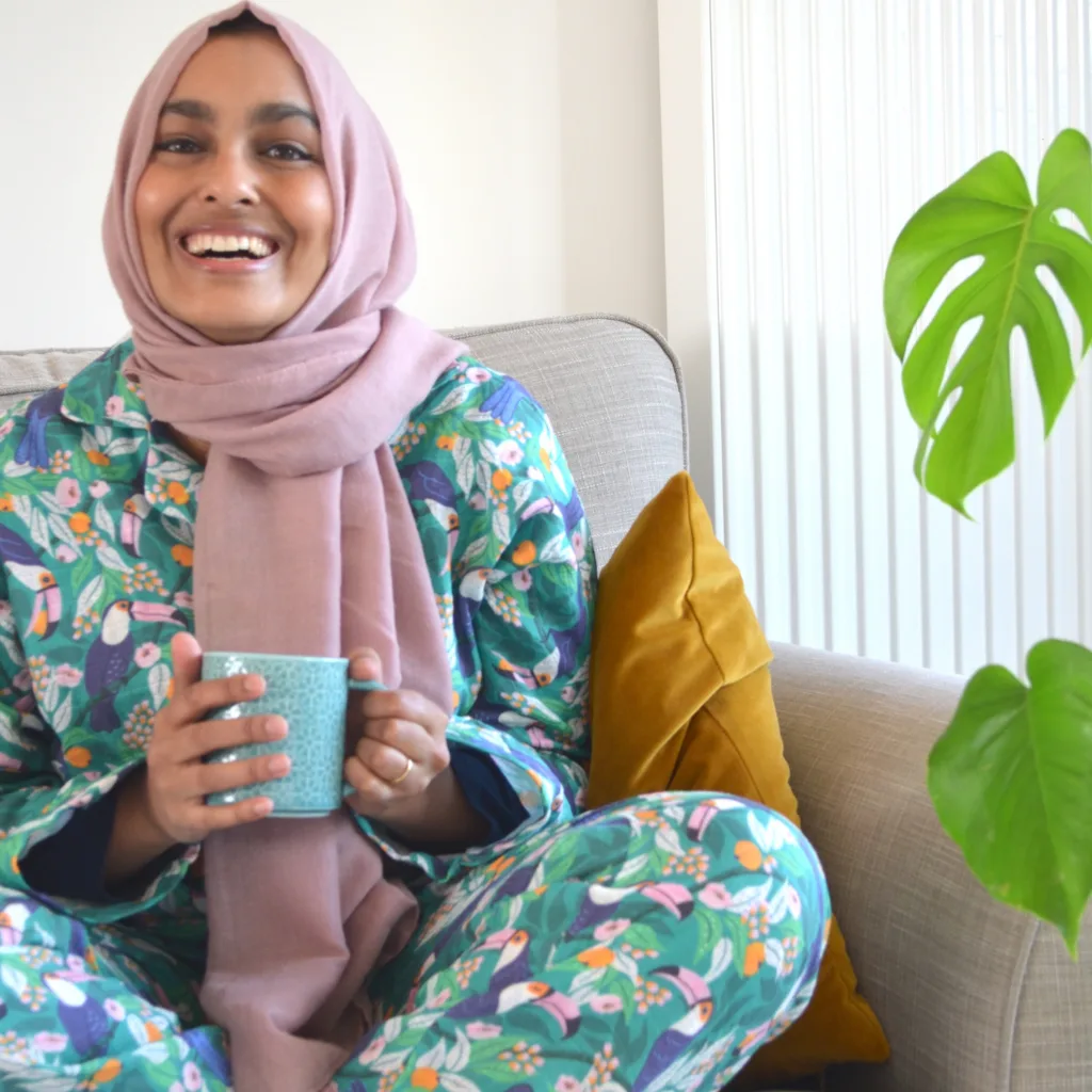 Rumana sits on a gray couch holding a mug in her hands, looking at the camera and smiling. She is wearing a dusty pink hijab and pajamas with a tropical print featuring toucans, pink and orange flowers and a bright blue background. To the right are a few
