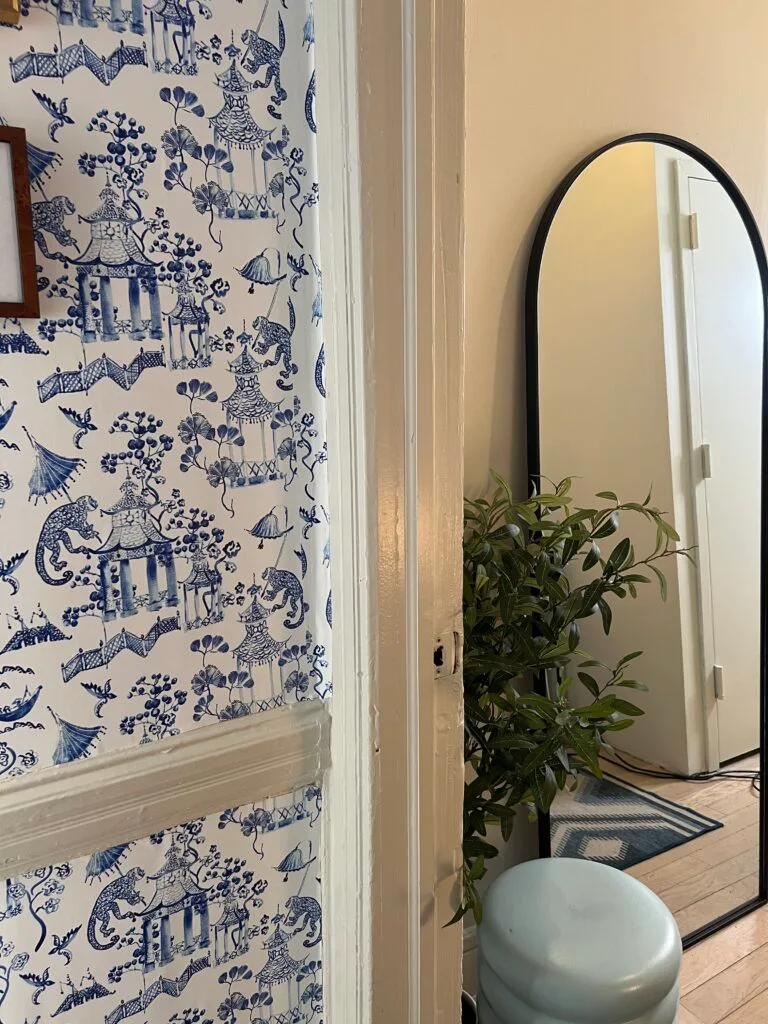 Image of a redecorated hallway with a blue-and-white chinoiserie design featuring wallpaper with a garden scene on both the walls. A mirror with a rounded top has been placed to the right side of the entrance to the hallway along with a plant and a small
