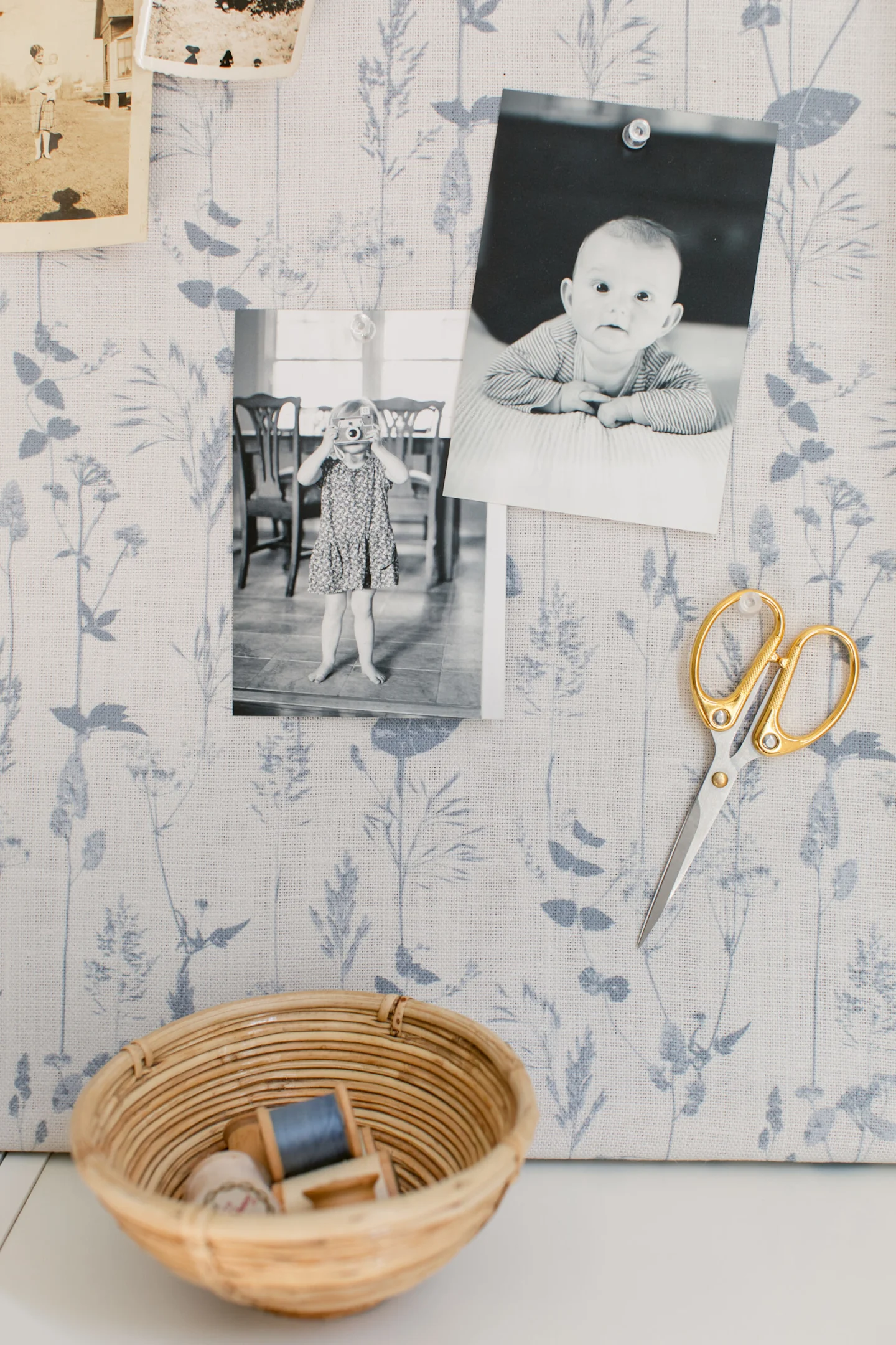  A close up of a fabric-covered bulletin board resting on the top of a low white bookshelf. The fabric design has a white background and light blue flowers. To the board’s bottom right are two black-and-white photos of small children, one of an infant loo