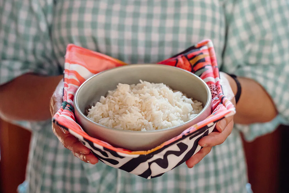 A close up of a person wearing a blue-and-white gingham dress holds a bowl of rice that itself is held in a fabric bowl cozy. The fabric on the top side of the cozy features bright flowers in inks, reds, yellows and white. The fabric on the bottom side fe