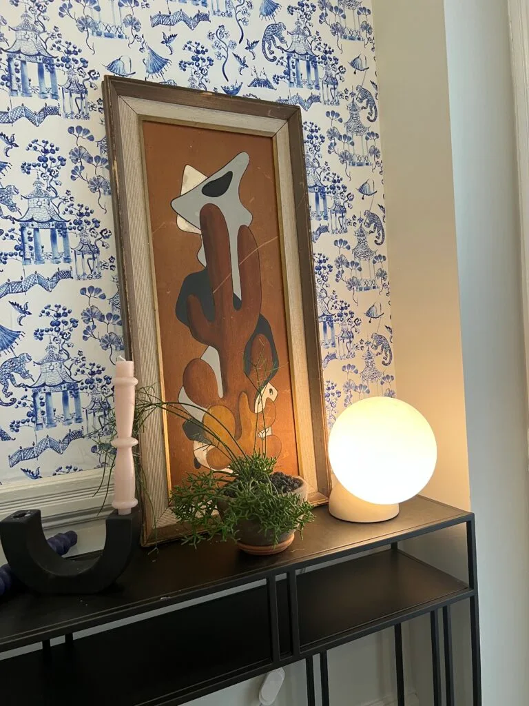Close up of a small black table with a round soft white lamp to the left in a redecorated hallway with a blue-and-white chinoiserie design featuring wallpaper with a garden scene on both the walls. A small plant in a terracotta planter and a pink candle i