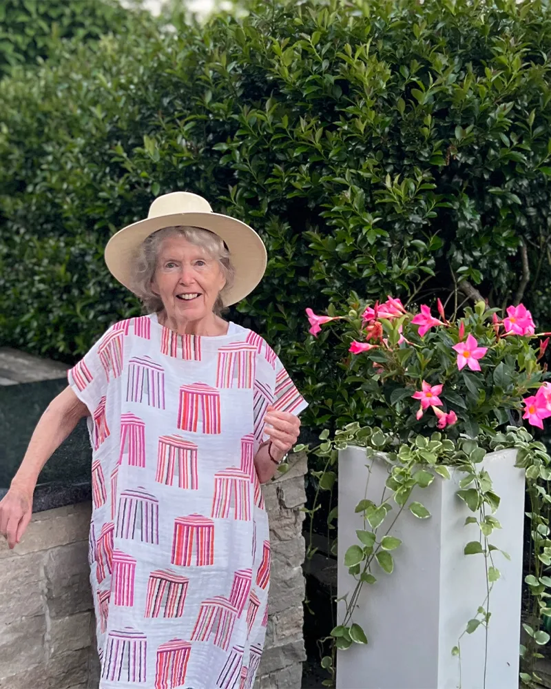 Danika's mom wears a caftan and stands next to some pink flowers. The caftan features hot pink and orange cabanas on a white background.