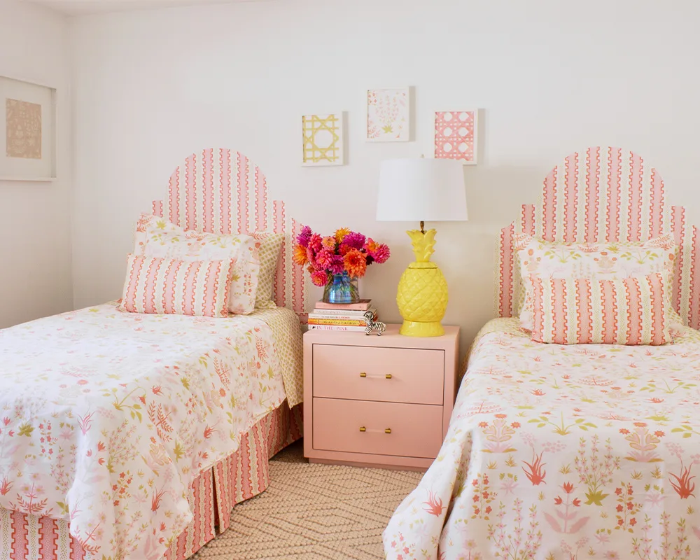 The finished room. The designs on the art, bedding and headboards are light pink and green florals. A pink bedside table with a bouquet of bright flowers to the left and a yellow lamp to the right is between the two beds.