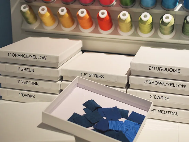 Small labeled white jewelry boxes rest on a white table. One box is open exposing small blue fabric squares. Different colored spools of thread are in the distance on a holder.