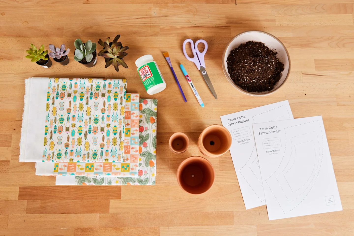 All the materials from the list below, which you’ll need for the fabric-covered flower pot tutorial, are laid out on a brown wooden table.