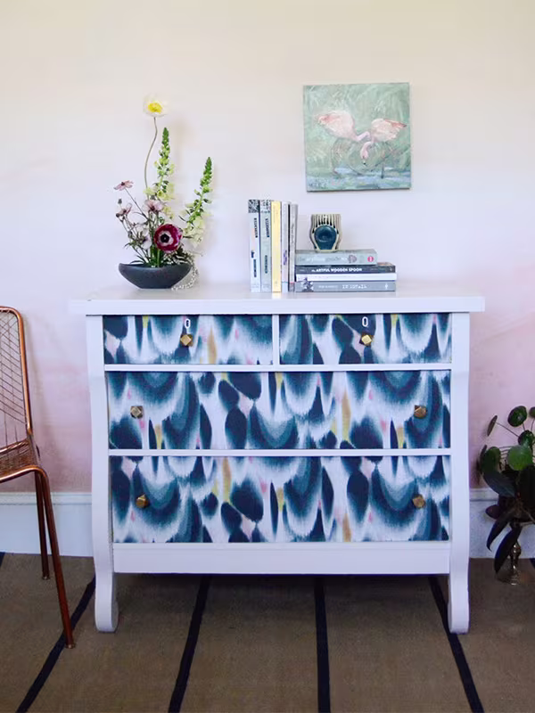 A white dresser with blue pink and gold wallaper sits in front of a ombred wall. A chair and plant sit on other side of a the dresser. A plant and stack of books rest on the the dresser.