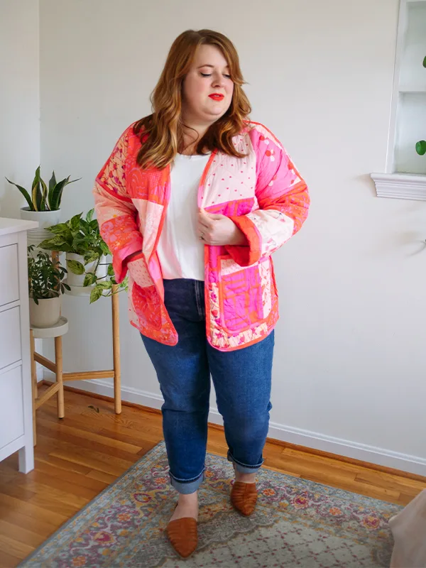 Meg wears her cheater quilt quilted jacket featuring pink prints and delicate florals. She is standing in front of a white wall.