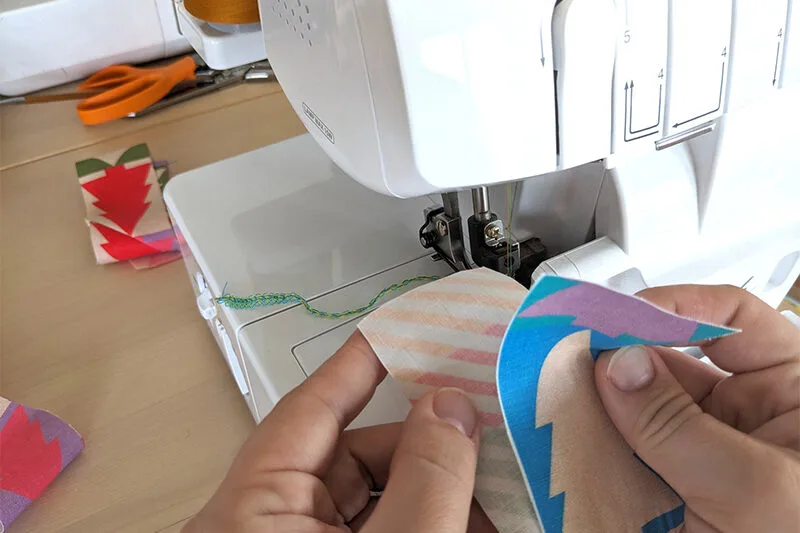 Two hands are lining up two 2”-wide strips of fabric at the tops, wrong sides together, in the middle of the image. The strips of fabric have a Christmas tree repeating design, also in jewel tones. A white sewing machine is in front of their hands.