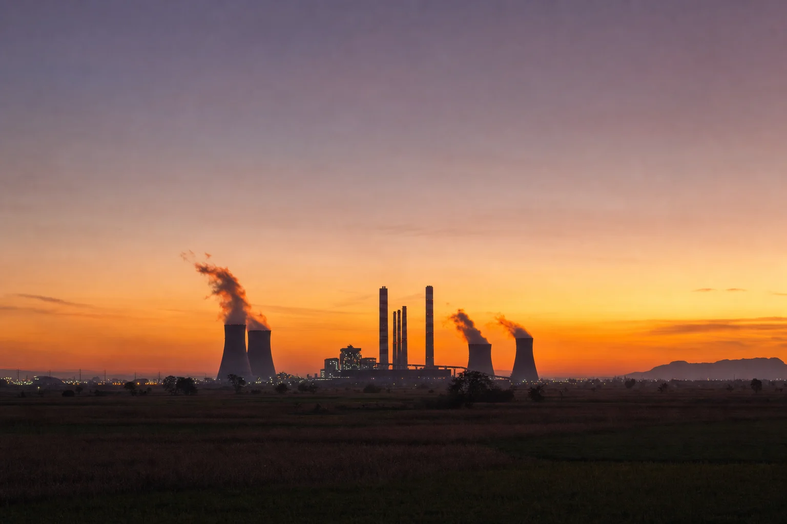 A coal-fired power station silhouetted at sunset across open fields, steam plumes lit orange from below as daylight drains away.