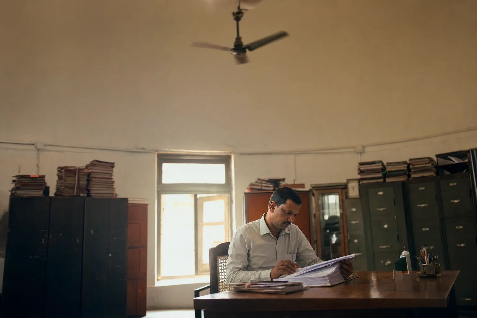 A utility procurement official studying a thick folder of papers at a wooden desk in a hot office, ceiling fan above and harsh daylight through a dusty window.