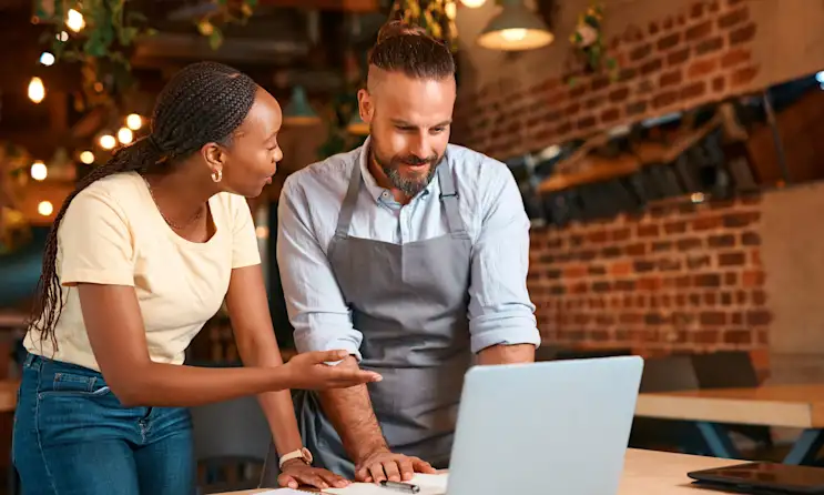 A woman and man stand over a computer in a restaurant and discuss performance