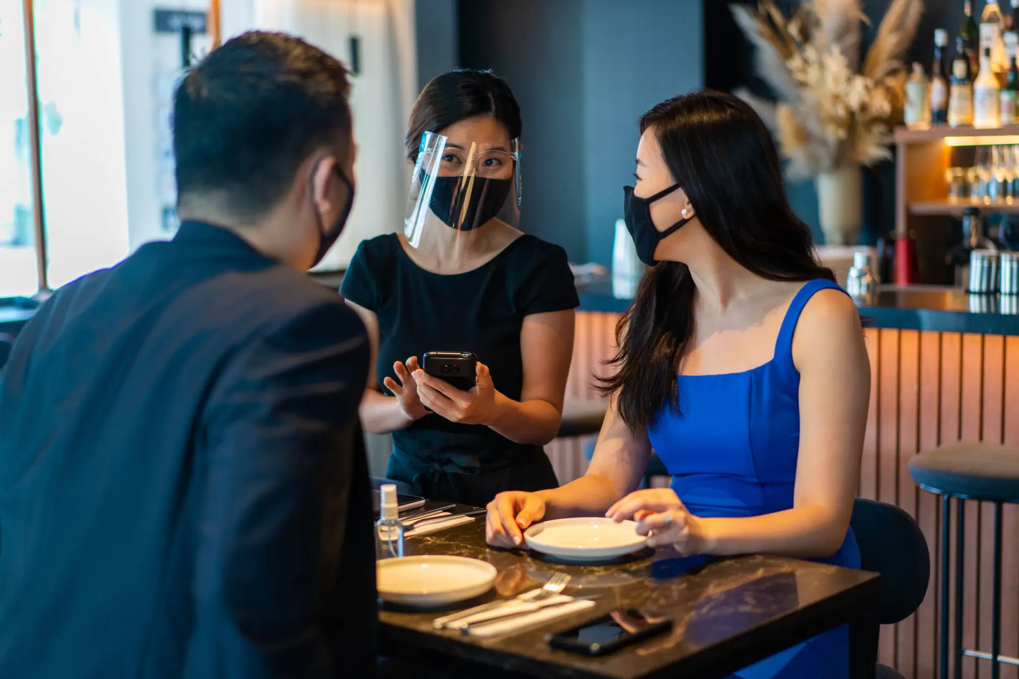 a man and a woman sitting at a table wearing face masks putting in a food order with a waitress wearing PPE