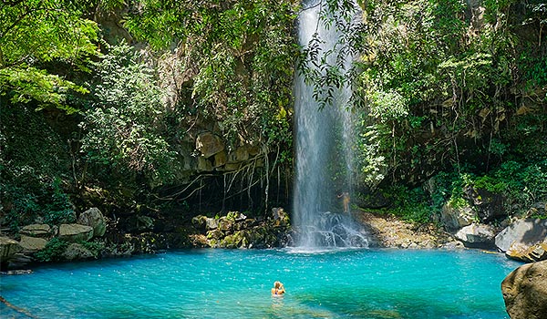 Femme nageant dans un bassin cristallin au pied d’une grande chute d’eau dans la forêt
