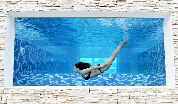 Un mur de verre à travers lequel on voit une femme qui nage dans la piscine Mermaid