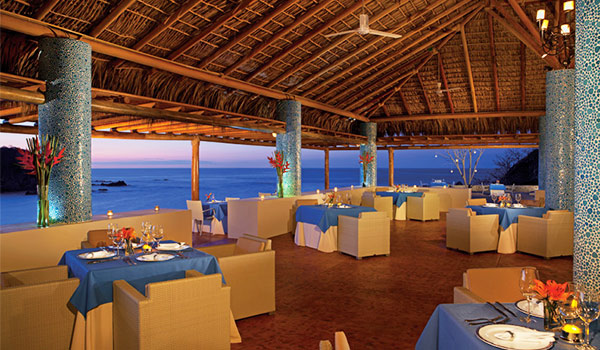 Élégant restaurant à aire ouverte doté d’un toit en chaume et donnant sur la mer au coucher de soleil