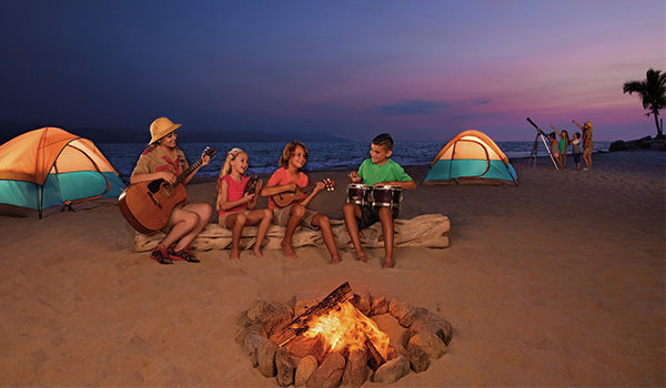 Kids sitting around a campfire on the beach with tents and a telescope in the background
