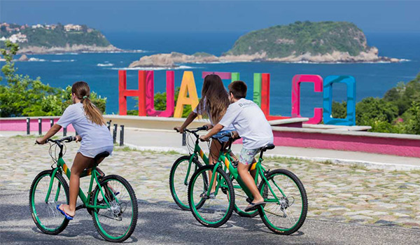 Three people riding bikes past a colourful Huatulco sign overlooking the coast