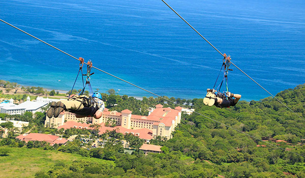 Two people on dual ziplines flying high above the coast