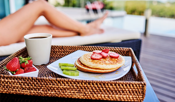 Pancakes, strawberries and a cup of coffee on a wicker tray next to a girl on a sun lounger