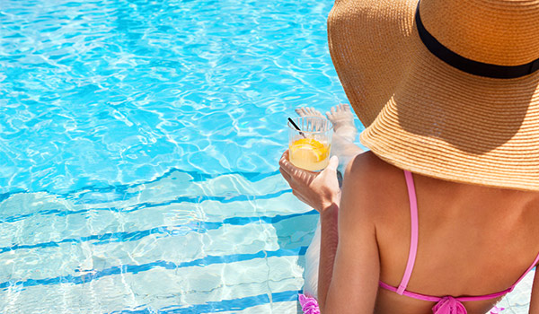 Glamorous woman sitting in a pool holding a cocktail and wearing a floppy straw hat