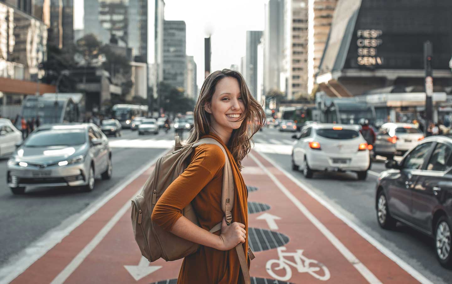 Woman wearing a backpack in the middle of the street, smiling. Image is courtesy of Guilherme Stecanella