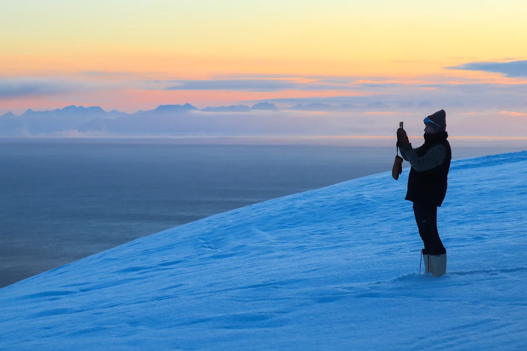 Woman stand in deep snow whilst taking a picture of the sunset during the Sunny Winter on Svalbard. Photo Credit: Eveline Lunde / Hurtigruten Svalbard