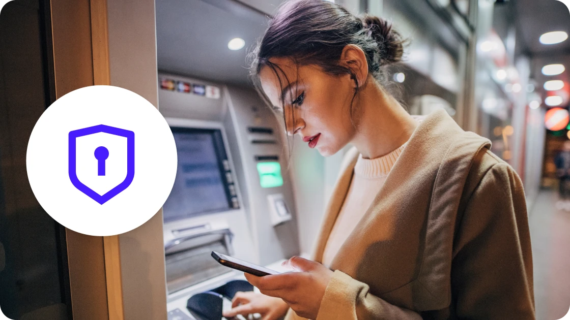 Photo of a woman checking her phone while standing at an ATM, with a shield and lock icon.