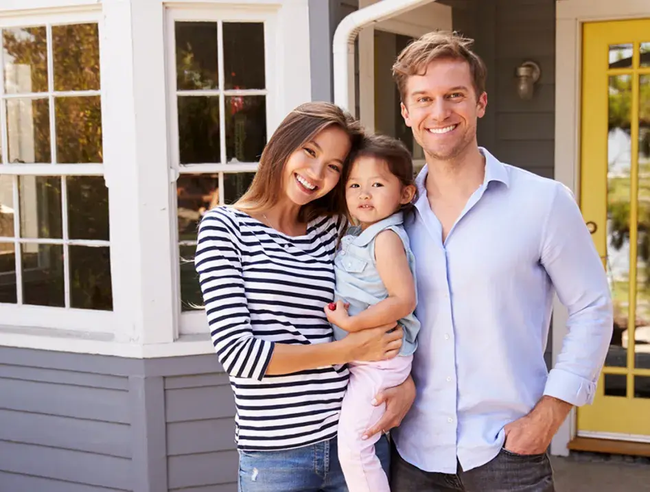 Photo of two adults and a young child smiling in front of a house.