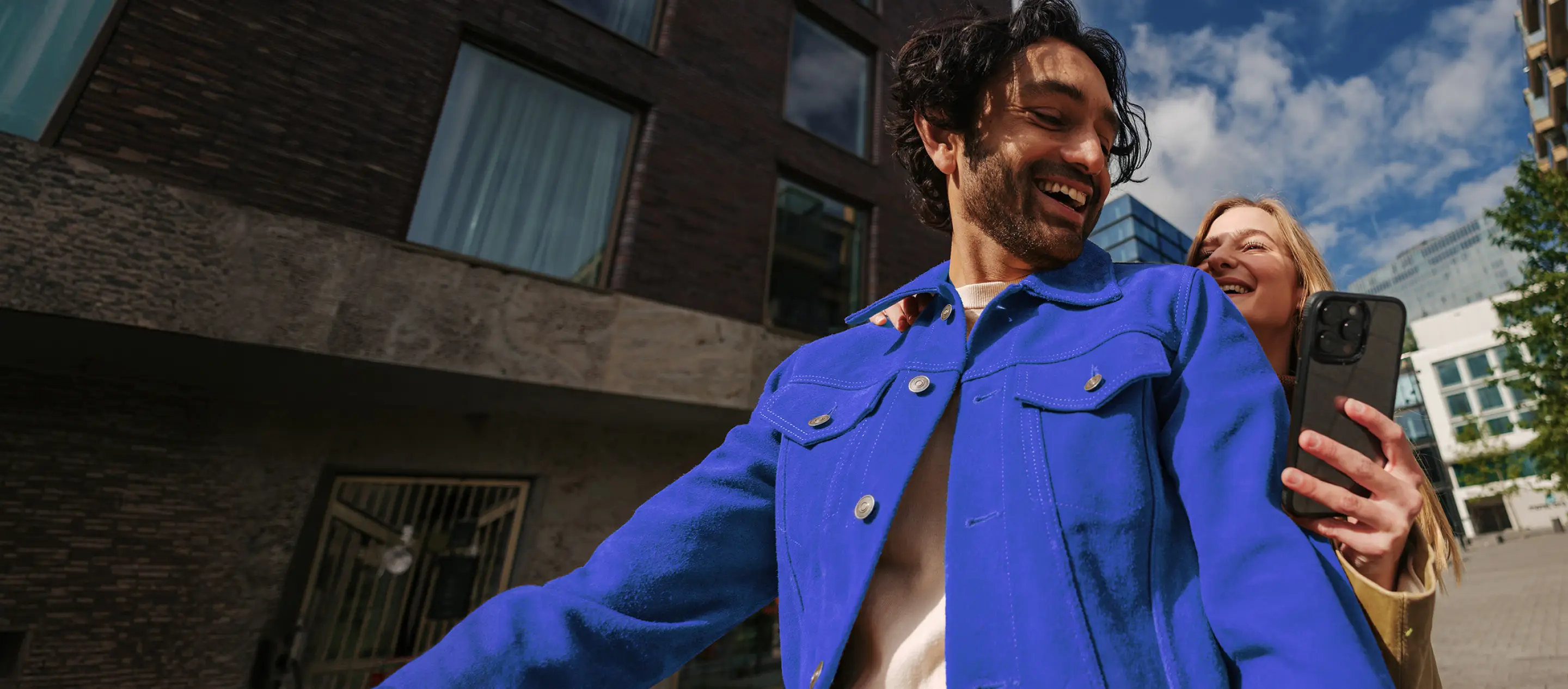 Photo of a man and a woman riding a bicycle together outdoors, both laughing. The man wears a blue jacket and steers the bike through a city street lined with buildings.