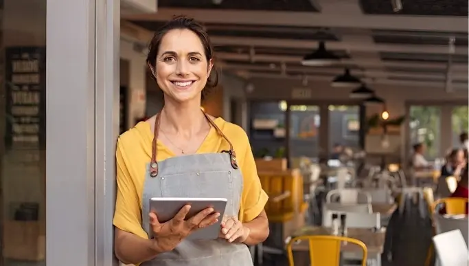 Woman in apron smiling while using tablet in the doorway of a restaurant