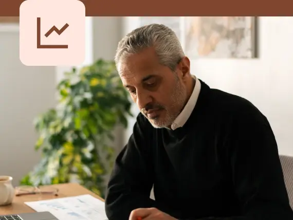 Photo of a man seated at a desk reviewing printed documents, with a line chart icon overlay in the upper left corner.