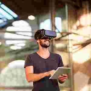 A happy man in a black shirt wears a virtual reality headset and holds a tablet.