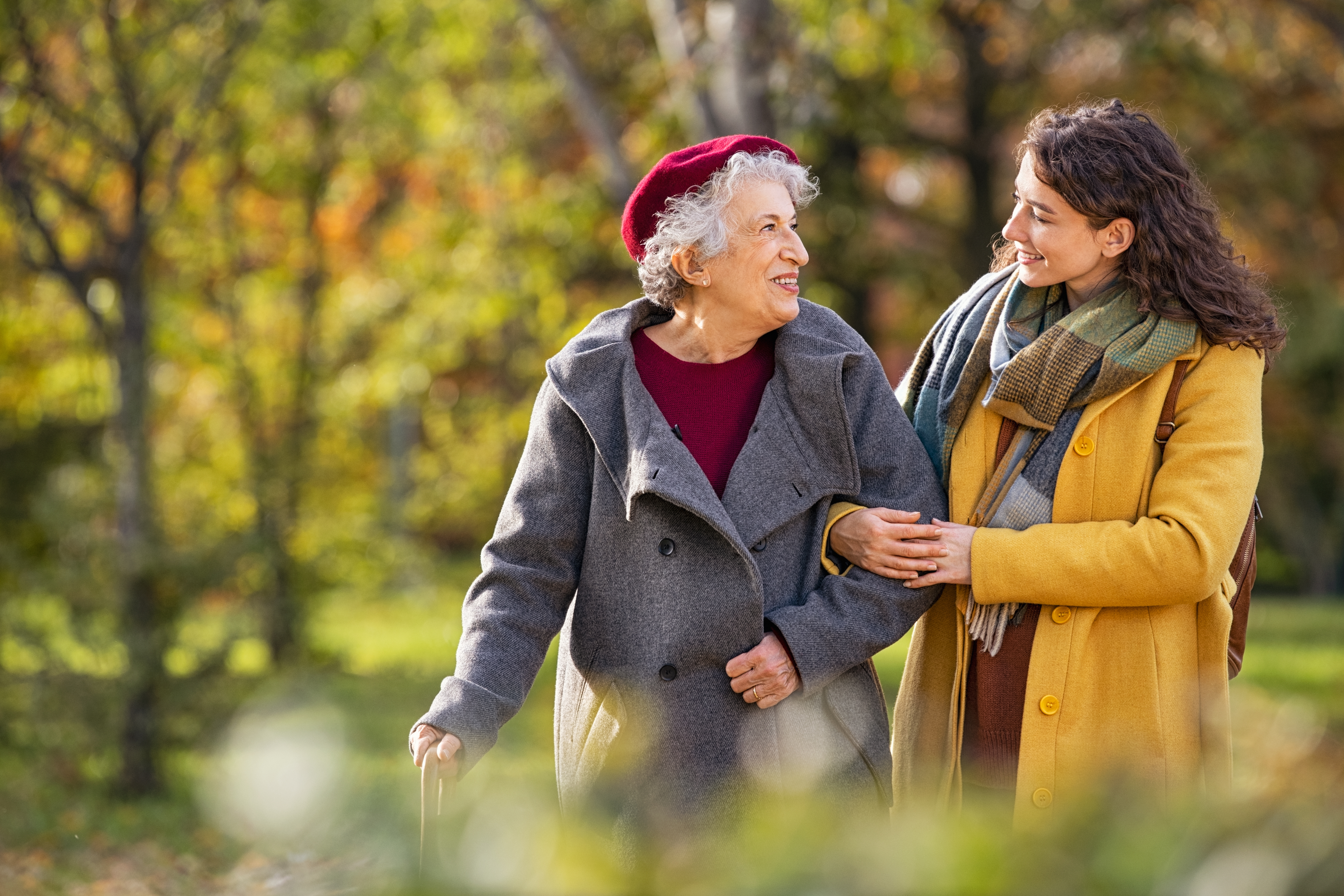 A young woman in a yellow coat assists a smiling elderly woman with a cane on an autumn park walk.