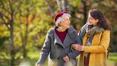 A young woman in a yellow coat assists a smiling elderly woman with a cane on an autumn park walk.