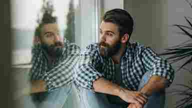 Young, pensive man in a checkered shirt sitting by the window and looking through it.