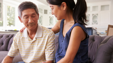 A woman is comforting an elderly man while sitting on the couch with her arms around his shoulder.