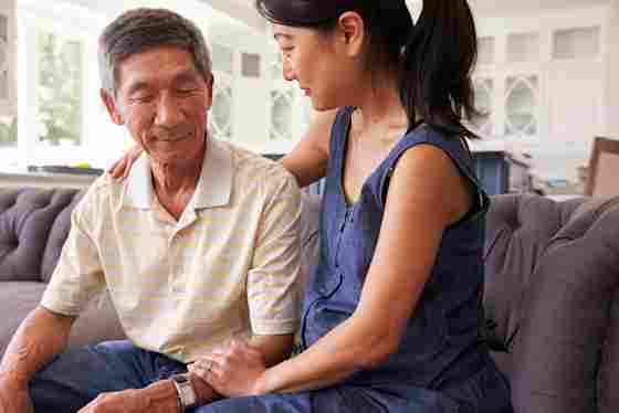 Woman is comforting an elderly man while sitting on the couch with her arms around his shoulder.