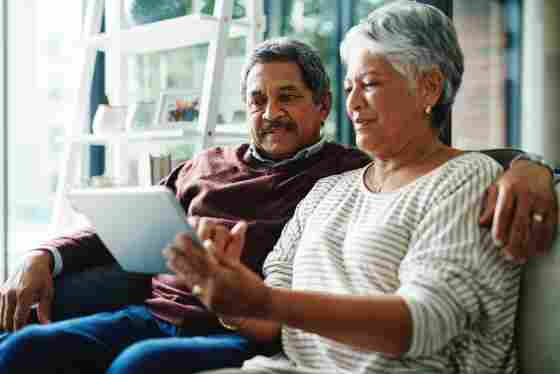 A mature senior older couple using a digital tablet while relaxing at home. The man has his arm around the woman as they both look at the tablet.