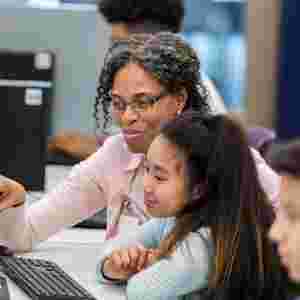 Teacher Helping Students Working At Computers In Classroom Pointing ad something on screen