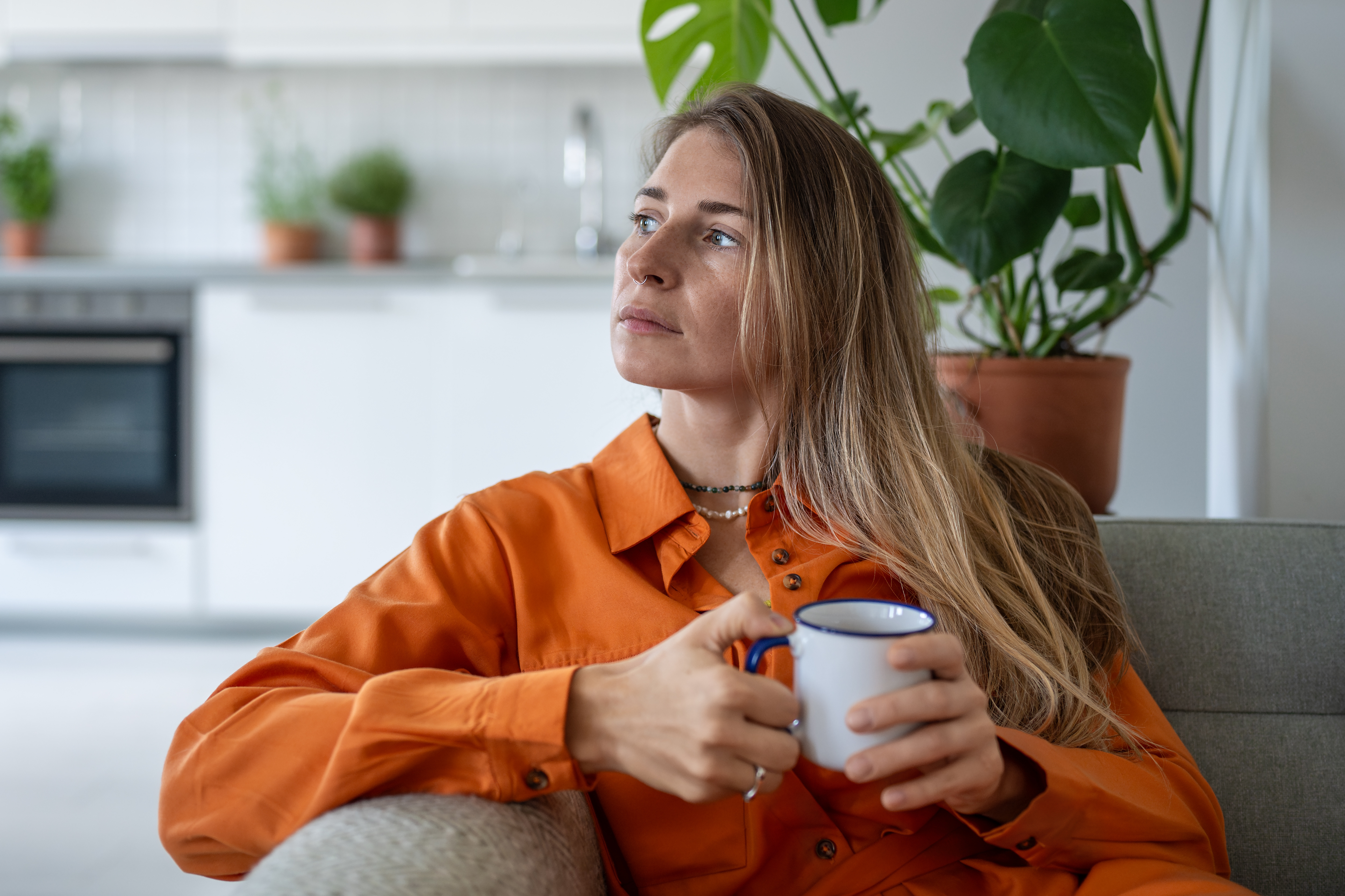 A thoughtful woman in an orange shirt holds a white mug while sitting on a sofa, with houseplants and a kitchen in the background.