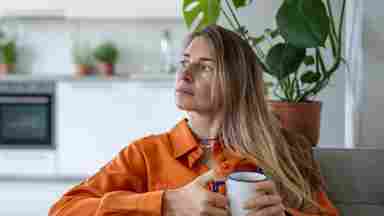 A thoughtful woman in an orange shirt holds a white mug while sitting on a sofa, with houseplants and a kitchen in the background.