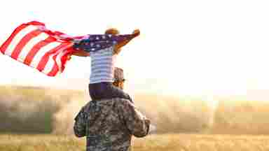 Rear view of a male soldier who is carrying a child holding an American flag on his shoulders. They are walking into a field on a sunny day.