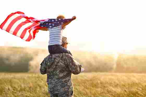 Rear view of soldier, carrying a child with an American flag and walking into a field on a sunny day.