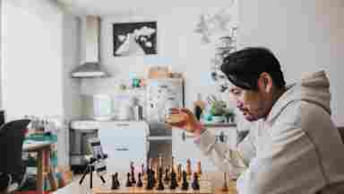 A man of color is sitting at a table and playing chess; he is holding a glass of wine and having a video call with a friend.