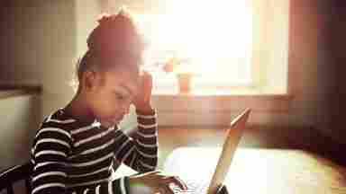 A side-view of a young Black girl working on her homework at home; she is typing information on a laptop as the sun glares brightly through the window. Used in Limitless brochure.