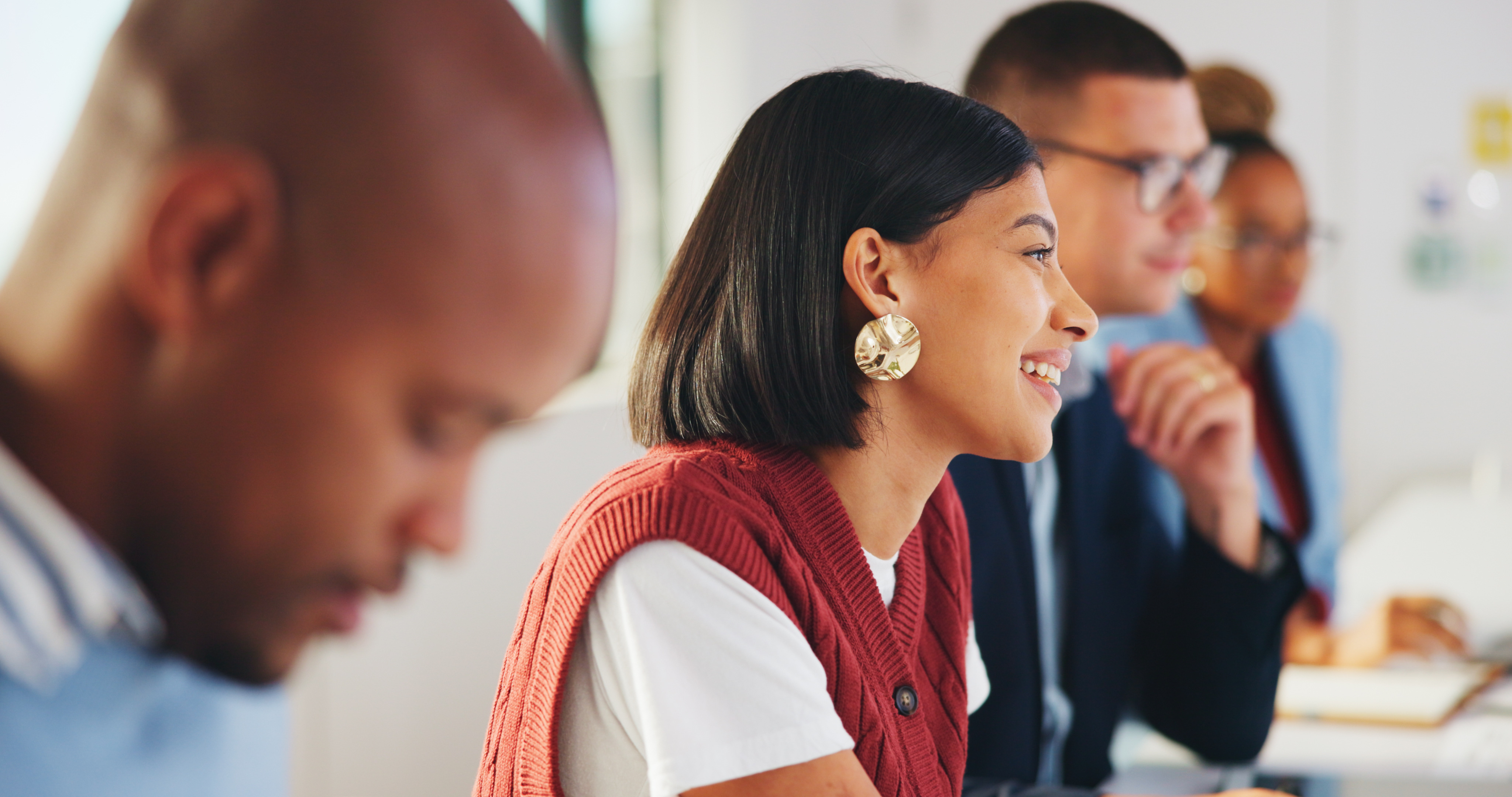 A smiling woman in a red vest sits among diverse colleagues during a meeting in a bright office setting.
