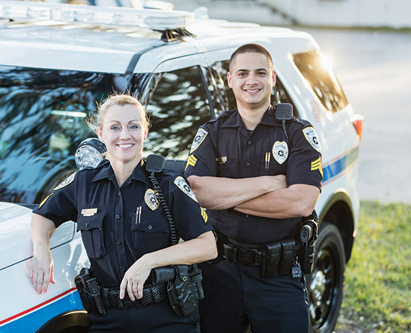 Two police officers in uniform stand in front of a police vehicle. The officer on the right has their arms crossed while the one on the left leans on the hood of the vehicle.