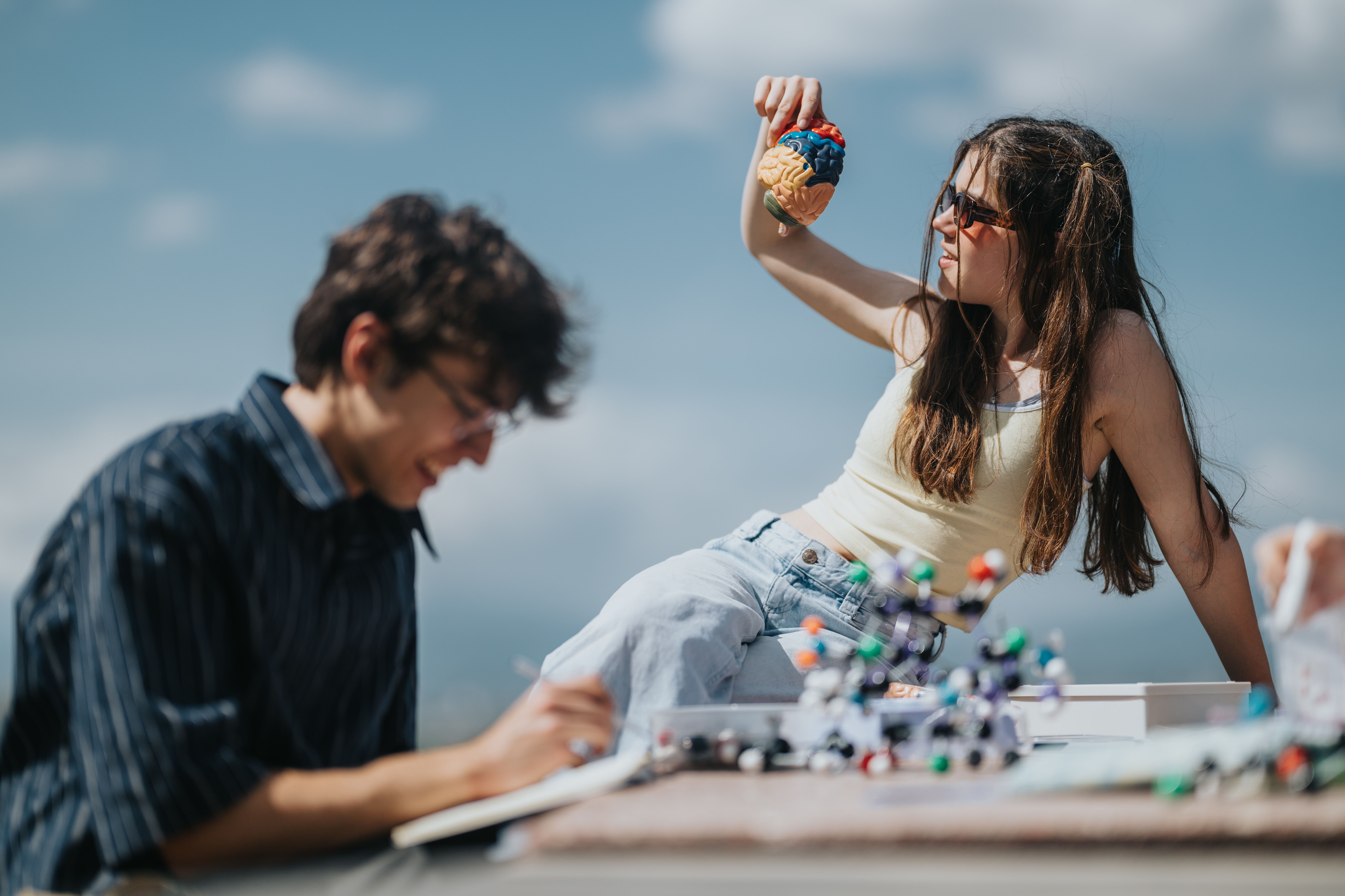 Two students study outdoors; a girl holds a colorful brain model while a boy writes notes, with molecular models on the table.