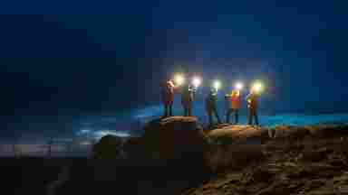 A group of night sky photographers stand on a large stone, each holding a light toward the night sky.